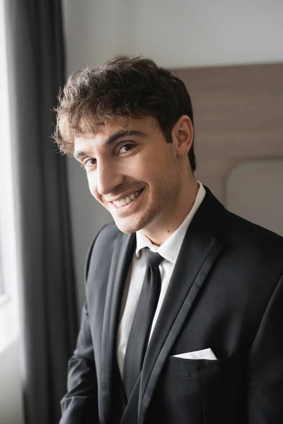 Porträt eines glücklichen Mannes in eleganter, formeller Kleidung mit schwarzer Krawatte und weißem Hemd, der im modernen Hotelzimmer in die Kamera blickt, Bräutigam am Hochzeitstag, besonderer Anlass — Stockfoto