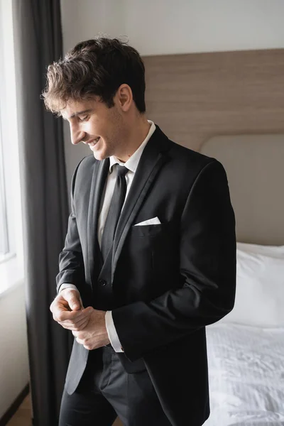 Portrait de jeune homme heureux en costume noir classique avec cravate et chemise blanche souriant tout en se tenant dans la chambre d'hôtel moderne, marié le jour du mariage, occasion spéciale — Photo de stock