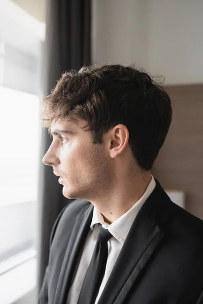Портрет хорошого чоловіка в стильному формальному одязі з чорною краваткою і білою сорочкою, що дивиться на вікно в сучасному готельному номері, нареченому на день весілля, особливий випадок, вид збоку — стокове фото