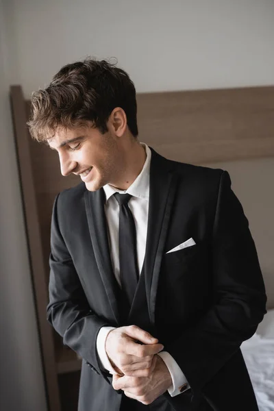 Novio positivo en ropa formal con elegante corbata negra y camisa blanca sonriendo y de pie en la habitación de hotel moderna, novio en el día de la boda, ocasión especial - foto de stock