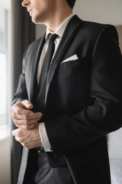 Обрізаний вид на нареченого в офіційному одязі з стильною чорною краваткою і білою сорочкою, що стоїть в сучасному готельному номері, регулюючи наручники, наречений на день весілля, особливий випадок — стокове фото