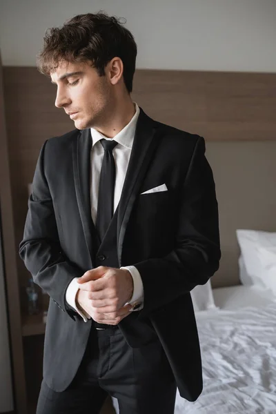 Beau marié en tenue formelle avec cravate noire chic et chemise blanche debout dans la chambre d'hôtel moderne tout en ajustant les menottes, homme le jour du mariage, occasion spéciale — Photo de stock