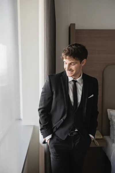 Glücklicher Bräutigam in eleganter formaler Kleidung mit schwarzer Krawatte und weißem Hemd, stehend mit Händen in Taschen auf Hosen in modernem Hotelzimmer am Fenster, Mann am Hochzeitstag, besonderer Anlass — Stockfoto