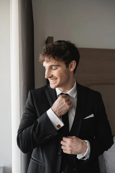 Happy and good looking man in classy formal wear with white shirt adjusting black tie while smiling in modern hotel room, groom on wedding day, special occasion — Stock Photo