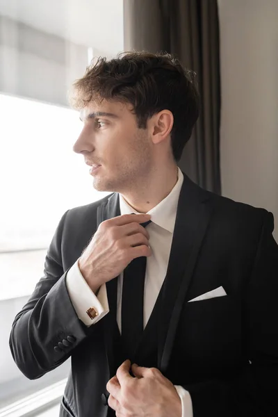 Novio de buen aspecto en ropa formal con clase con camisa blanca que ajusta la corbata negra mientras mira a la ventana en la habitación de hotel moderna, hombre en el día de la boda, preparándose para una ocasión especial — Stock Photo