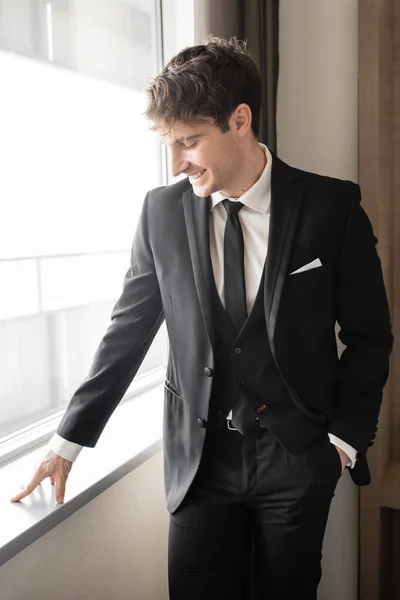 Glücklicher Mann in eleganter formaler Kleidung mit schwarzer Krawatte und weißem Hemd, der mit der Hand in der Tasche auf der Fensterbank in einem modernen Hotelzimmer neben dem Fenster steht, Bräutigam am Hochzeitstag, besonderer Anlass — Stockfoto
