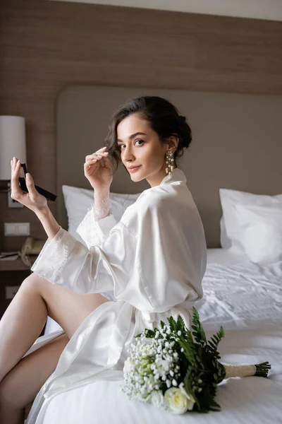 Привлекательная женщина в белом шелковом халате готовится к свадьбе, делая макияж и держа карманное зеркало, сидя на кровати возле свадебного букета в гостиничном номере, особый случай, молодая невеста — стоковое фото