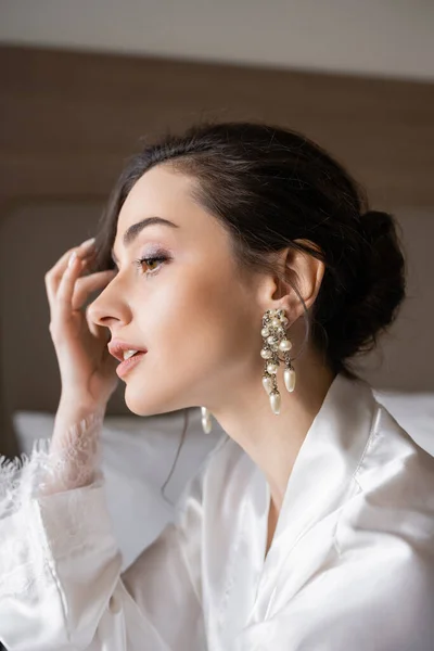 Porträt einer attraktiven Frau mit brünetten Haaren in weißem Seidenmantel, Perlenohrringen und makellosem Make-up, die sich auf ihre Hochzeit im Hotelzimmer vorbereitet, besonderer Anlass, junge Braut — Stockfoto