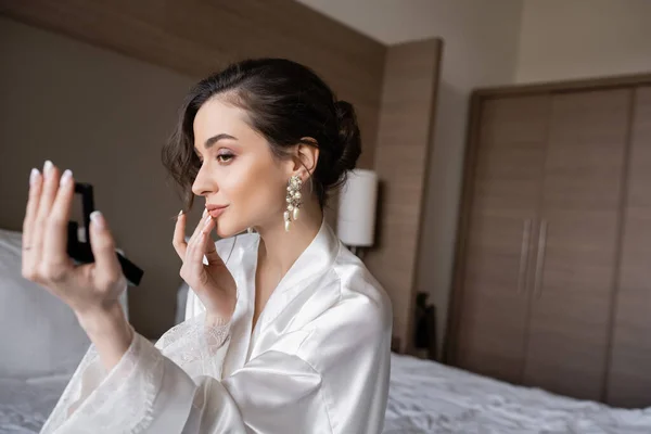 Привлекательная женщина в белом шелковом халате готовится к свадьбе, прикасаясь к губам, держа карманное зеркало и сидя на кровати в гостиничном номере в день свадьбы, особый случай, молодая невеста — стоковое фото