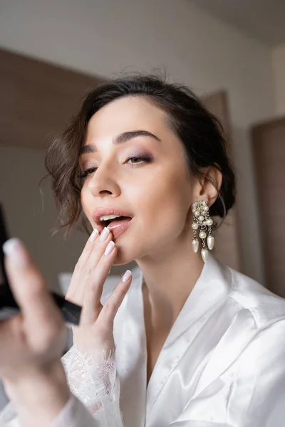 Young bride with brunette hair in white silk robe preparing for her wedding while touching lips, holding pocket mirror in hotel room on wedding day, special occasion, — Stock Photo