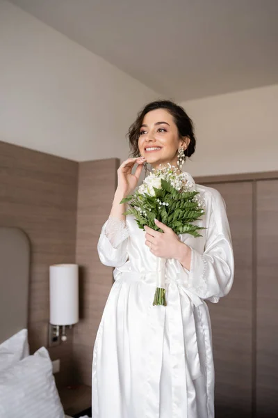 Young woman with brunette hair in white silk robe and pearl earrings holding bridal bouquet while preparing for her wedding in hotel room, special occasion, happy bride — Stock Photo