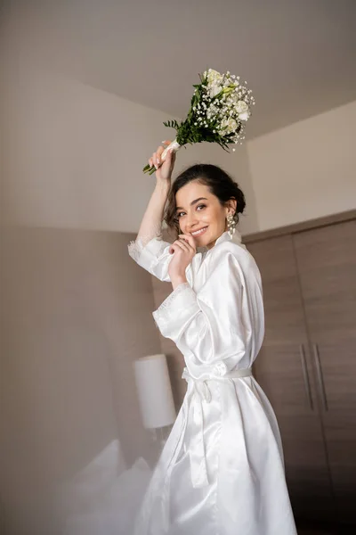 Alluring young woman with brunette hair in white silk robe and pearl earrings holding bridal bouquet while preparing for her wedding in hotel room, special occasion, happy bride — Stock Photo