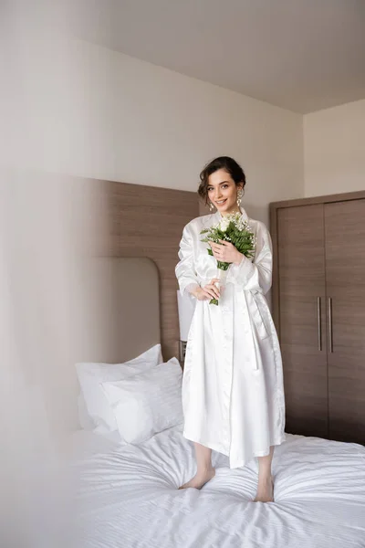 Jovem noiva alegre com cabelo morena em roupão de seda branca e brincos de pérola segurando buquê de noiva enquanto estava de pé na cama no quarto do hotel, ocasião especial, mulher feliz — Fotografia de Stock