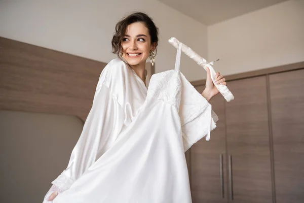 Happy young bride with brunette hair in white silk robe holding soft hanger with elegant wedding dress and smiling in bedroom of hotel room, special occasion, charming woman — Stock Photo
