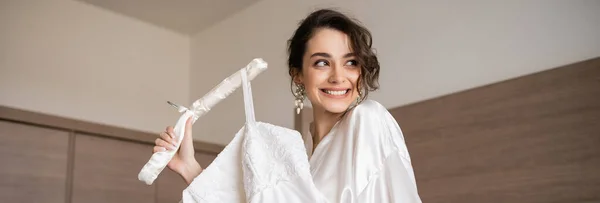 Happy young bride with brunette hair in white silk robe holding soft hanger with elegant wedding dress and smiling in bedroom of hotel room, special occasion, charming woman, banner — Stock Photo