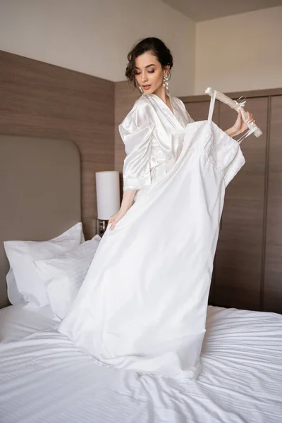 Enchanting young bride with brunette hair in white silk robe holding soft hanger with elegant wedding dress and standing on bed of hotel room, special occasion, charming woman — Stock Photo