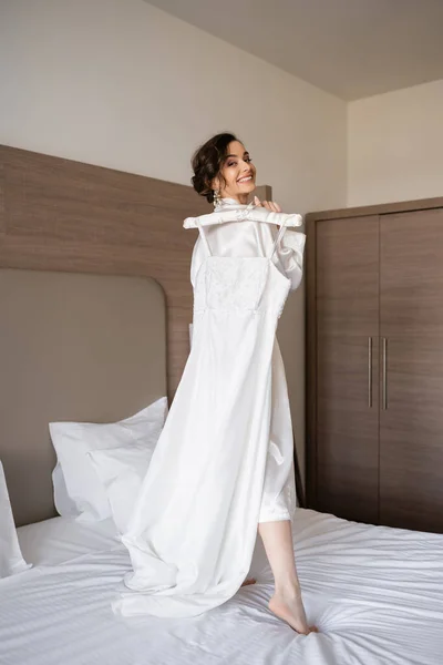 Noiva jovem feliz com cabelo morena em robe de seda branca segurando cabide macio com vestido de noiva elegante e de pé na cama no quarto do quarto de hotel, ocasião especial, mulher bonita — Fotografia de Stock