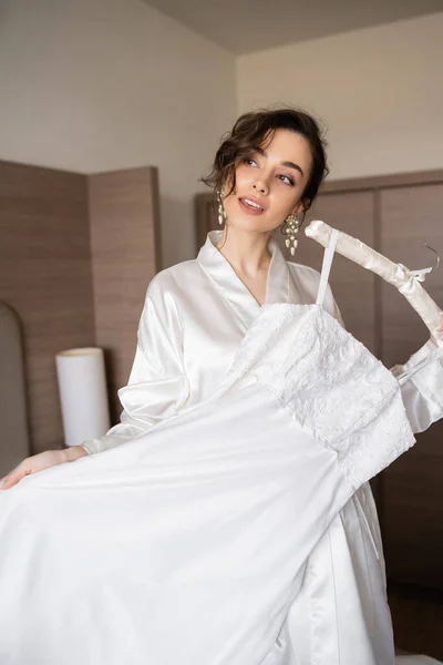 Young bride with brunette hair in white silk robe holding soft hanger with elegant wedding dress in bedroom of hotel room, special occasion, charming woman — Stock Photo