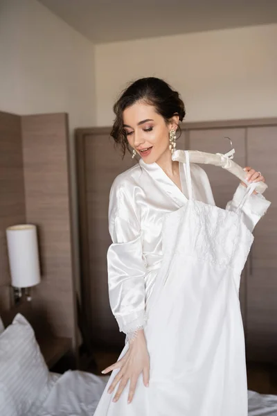 Amazed young bride with brunette hair in white silk robe holding soft hanger with elegant wedding dress in bedroom of hotel room, special occasion — Stock Photo