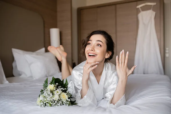Mulher feliz com cabelo morena deitado robe de seda branca e mostrando anel de noivado no dedo ao lado do buquê de noiva na cama no quarto do hotel com vestido de noiva no fundo borrado, jovem noiva — Fotografia de Stock