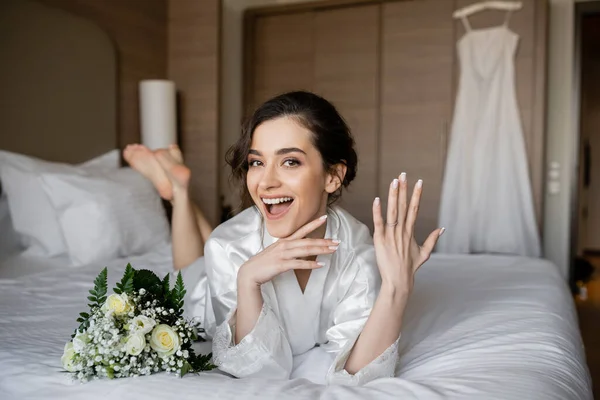 Mulher espantada com cabelo morena deitado robe de seda branca e mostrando anel de noivado no dedo ao lado do buquê de noiva na cama no quarto do hotel com vestido de noiva no fundo borrado, jovem noiva — Fotografia de Stock