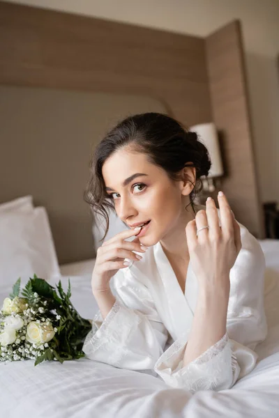 Charming woman with brunette hair lying in white silk robe and showing engagement ring on finger next to bridal bouquet in hotel room on wedding day, special occasion, young bride — Stock Photo