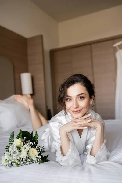 Encantadora mujer con el pelo moreno acostado en túnica de seda blanca con las manos apretadas junto al ramo de novia en la cama en la habitación de hotel el día de la boda, ocasión especial, novia joven - foto de stock