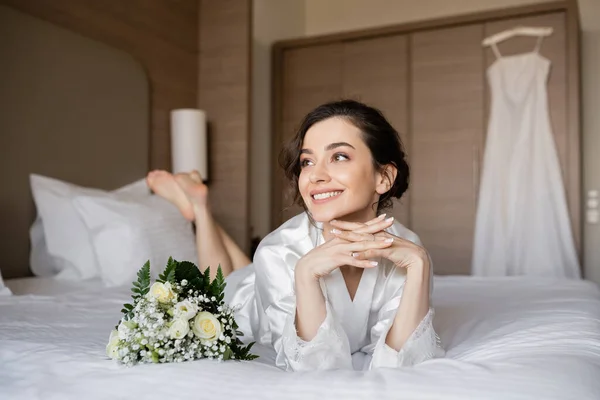 Femme gaie avec des cheveux bruns couché en robe de soie blanche avec les mains serrées à côté du bouquet de mariée sur le lit dans la chambre d'hôtel avec robe de mariée sur fond flou, occasion spéciale, jeune mariée — Photo de stock
