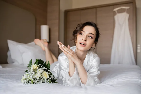 Великолепная женщина с брюнеткой волосы и обручальное кольцо на пальце глядя в сторону и лежа в белом шелковом халате рядом с свадебный букет на кровати с свадебным платьем на размытом фоне, молодая невеста — стоковое фото