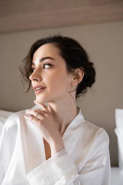 Young woman with brunette hair in white silk robe showing engagement ring on finger while looking away in hotel room on wedding day, special occasion, young bride — Stock Photo