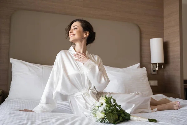 Jovem alegre com cabelo morena e anel de noivado no dedo sentado em robe de seda branca perto de buquê de noiva na cama no quarto do hotel no dia do casamento, ocasião especial, jovem noiva — Fotografia de Stock