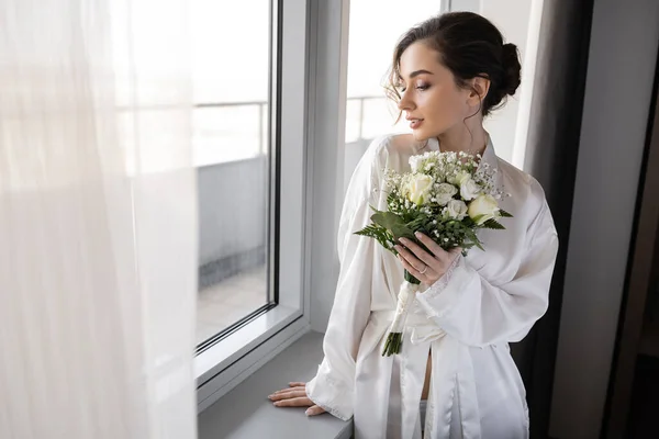 Jeune femme avec bague de fiançailles sur le doigt debout en robe de soie blanche et tenant bouquet nuptiale à côté du rideau de tulle et fenêtre dans la suite de l'hôtel, occasion spéciale, mariée le jour du mariage — Photo de stock