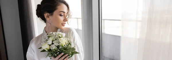Молодая женщина с брюнетками, стоящая в белом шелковом халате и держащая свадебный букет рядом с тюлем занавес и окно в номере отеля, особый случай, невеста в день свадьбы, баннер — стоковое фото