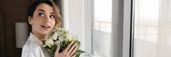 Mulher espantada com anel de noivado no dedo de pé em roupão de seda branca e segurando buquê de noiva enquanto olha para cima ao lado da janela na suíte do hotel, ocasião especial, noiva no dia do casamento, banner — Fotografia de Stock
