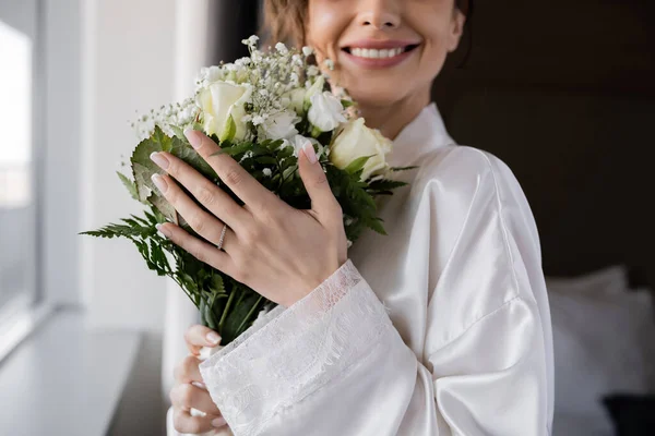 Обрезанный вид счастливой невесты с обручальным кольцом на пальце стоя в белом шелковом халате и держа свадебный букет рядом с окном в номере отеля, особый случай — стоковое фото