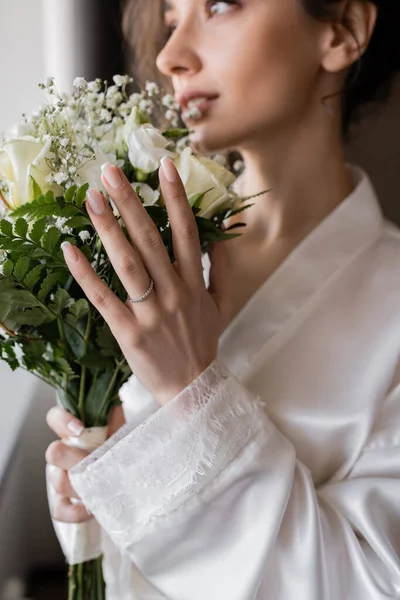 Blurred bride with engagement ring on finger standing in white silk robe and holding bridal bouquet in modern hotel suite on wedding day, special occasion — Stock Photo
