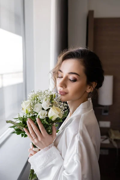 Молодая женщина с обручальным кольцом на пальце стоя в белом шелковом халате и глядя на цветочный букет невесты рядом с окном в номере отеля, особый случай, невеста в день свадьбы — стоковое фото