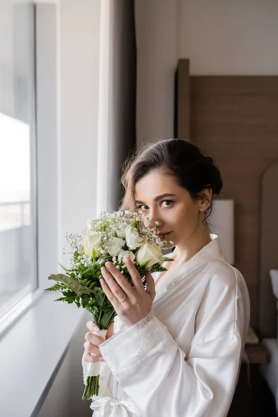 Young bride with engagement ring on finger standing in white silk robe and smelling bridal bouquet next to window in hotel suite, special occasion, wedding day — Stock Photo