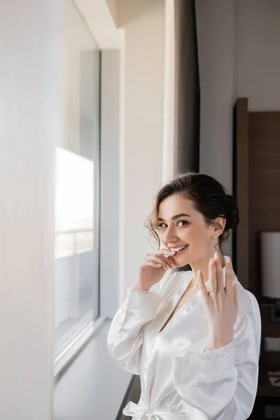 Excited young bride with brunette hair in white silk robe showing engagement ring on finger and smiling next to window in hotel suit on wedding day, special occasion, hand near lips — Stock Photo