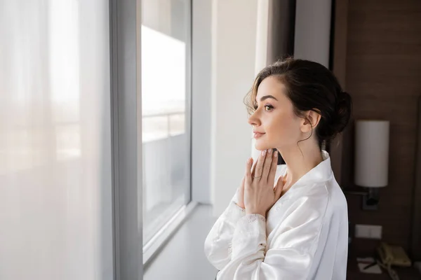 Side view of hopeful young woman with engagement ring on finger standing in white silk robe with praying hands and looking through window in hotel suite, special occasion, bride on wedding day — Stock Photo