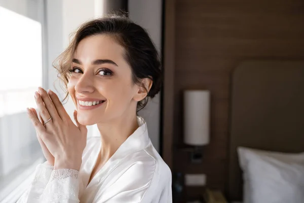 Portrait of delightful young woman with engagement ring on finger standing in white silk robe and looking at camera in modern hotel suite, special occasion, bride on wedding day — Stock Photo