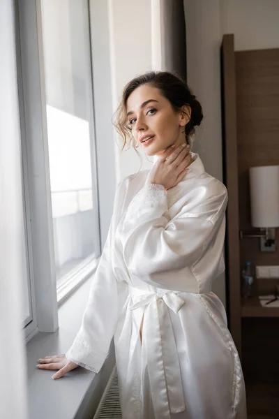 Молодая женщина с обручальным кольцом на пальце стоя в белом шелковом халате и глядя на камеру рядом с окном в номере отеля, особый случай, невеста в день свадьбы — стоковое фото
