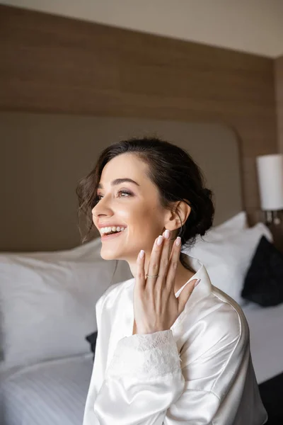 Excited young woman with brunette hair in white silk robe showing engagement ring on finger and smiling in hotel room on wedding day, special occasion, young bride — Stock Photo