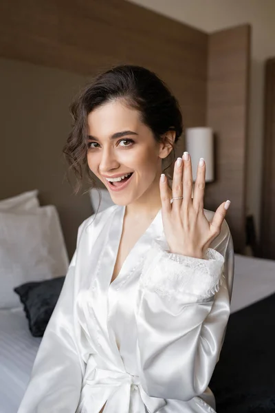 Novia joven asombrada con el pelo morena y la boca abierta posando en bata de seda blanca y mostrando anillo de compromiso en el dedo en la habitación de hotel el día de la boda, ocasión especial, mujer feliz - foto de stock