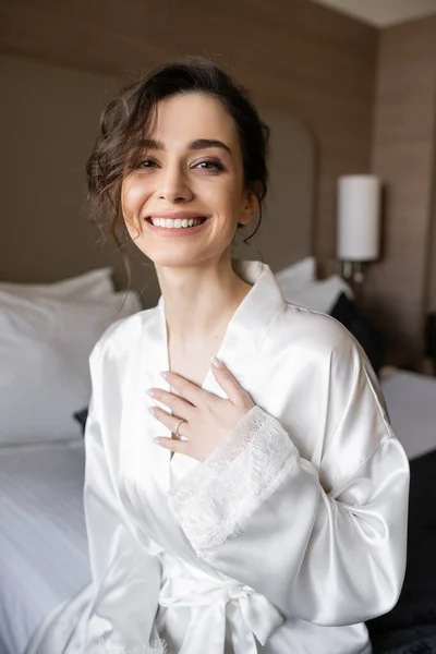 Happy young bride with brunette hair and engagement ring on finger smiling while sitting in white silk robe and looking at camera in hotel room on wedding day, special occasion — Stock Photo