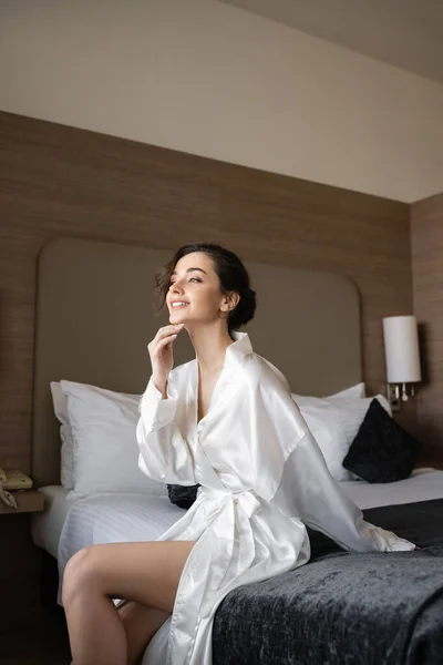 Happy young bride with brunette hair sitting in white silk robe on comfortable bed and looking away in hotel suite on wedding day, special occasion, young bride — Stock Photo