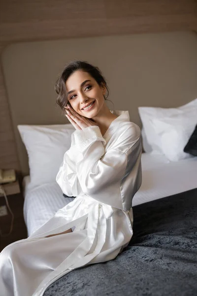 Novia encantadora y sonriente con cabello moreno sentado en bata de seda blanca en la cama cómoda con manta negra y mirando hacia otro lado en la suite del hotel el día de la boda, ocasión especial - foto de stock