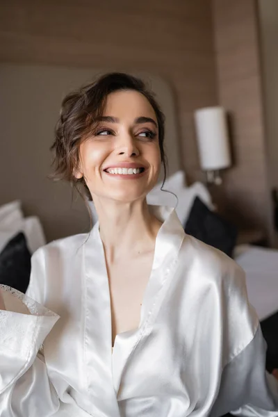Восхитительная женщина с брюнетками и свадебный макияж в белом шелковом халате улыбается и смотрит прочь в номере отеля в день свадьбы, особый случай — стоковое фото