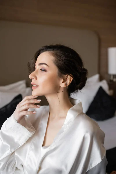 Side view of young bride with brunette hair and bridal makeup sitting in white silk robe on comfortable bed and looking away in hotel suite on wedding day, special occasion — Stock Photo