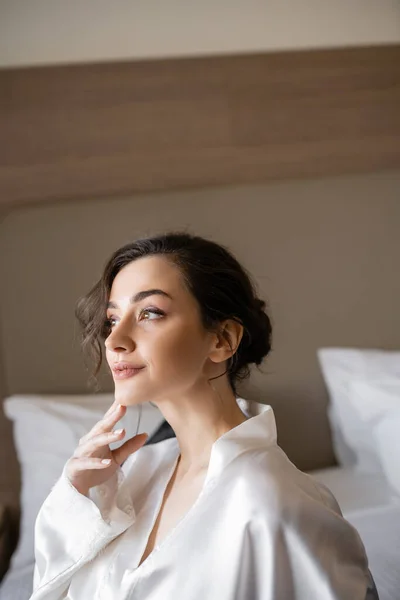 Alluring bride with brunette hair holding hand near face and sitting in white silk robe on comfortable bed and looking away in hotel suite on wedding day, special occasion — Stock Photo